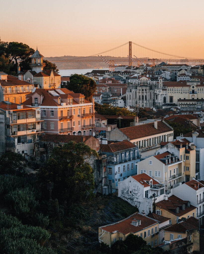 Panoramic view of Lisbon's colorful rooftops and historic buildings with the iconic 25 de Abril Bridge in the background during sunset.