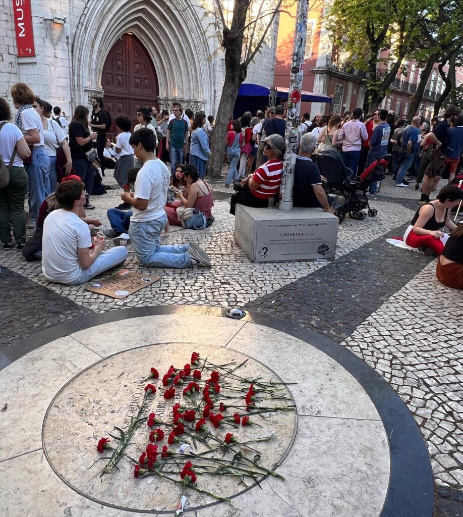 Lisbon in April: The Perfect Time To Visit 3 Crowds gather at the Lisbon memorial with red roses laid on the ground, reflecting a moment of remembrance in a lively city square during spring.