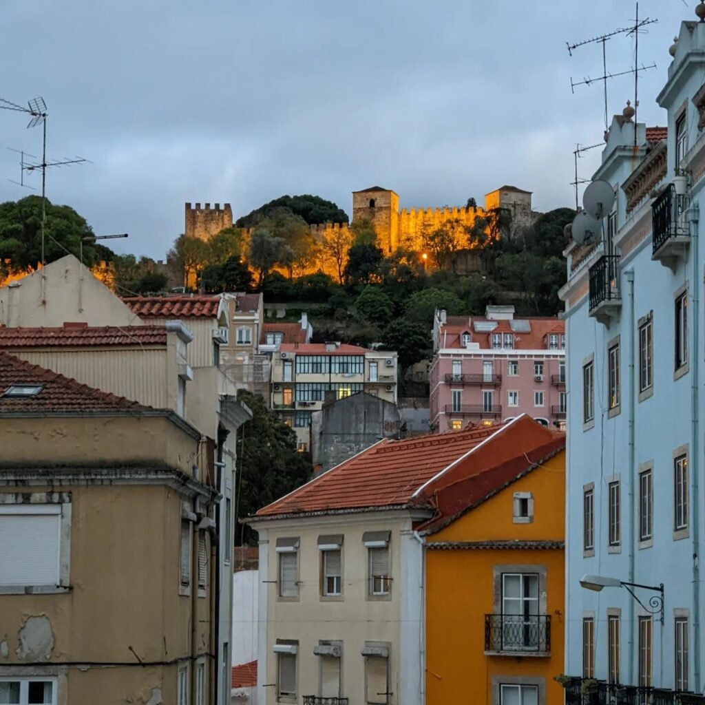 Scenic view of Lisbon's historic castle illuminated at sunset, overlooking colorful city buildings and the vibrant neighborhood below.