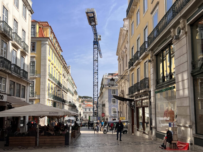 Busy Lisbon street scene featuring historic buildings, outdoor cafes, and a construction crane overhead, capturing the vibrant urban atmosphere of Lisbon.