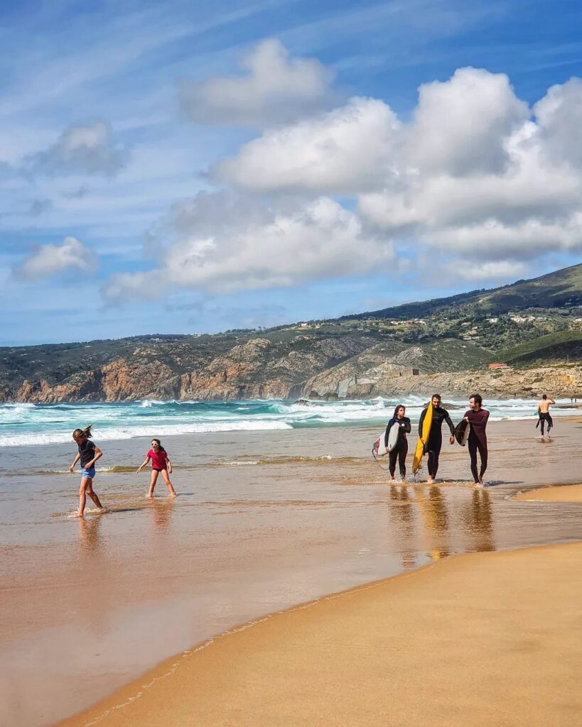 12 Best Beaches in Lisbon (Tourism Guide) 8 Scenic Lisbon beach scene featuring families and friends relaxing and walking along the sandy shore under a partly cloudy sky.