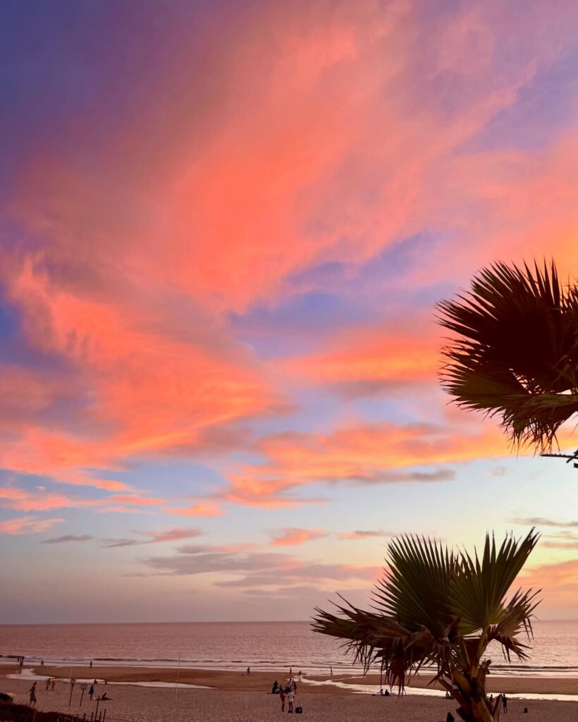 12 Best Beaches in Lisbon (Tourism Guide) 12 Sunset over Lisbon's beaches showcasing vibrant pink and orange clouds, palm trees, and calm ocean waters, perfect for relaxing seaside visits in Lisbon.