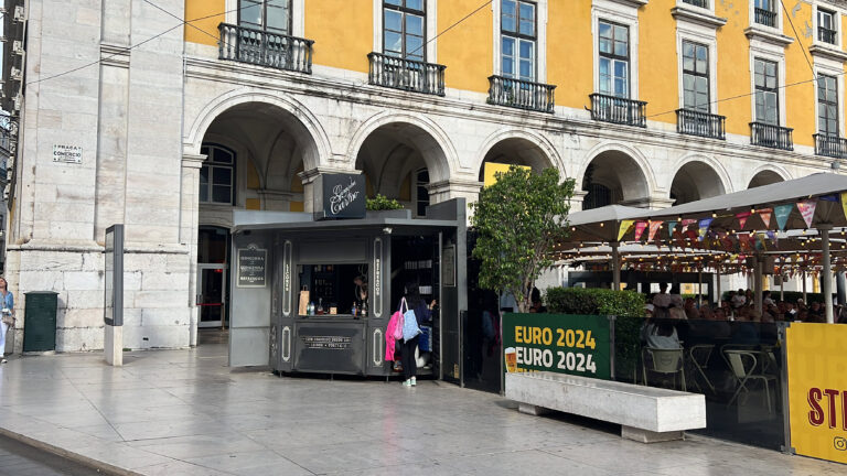 Historic Lisbon building with vibrant yellow facade, lively outdoor café area, and a city kiosk promoting EURO 2024, capturing the essence of LisboaVibes and Lisbon's bustling urban atmosphere.