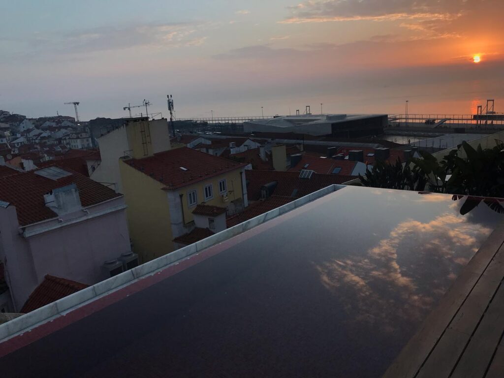 Alfama in Lisbon: A Tourist Guide 22 Scenic sunset view over Alfama district in Lisbon from a rooftop terrace, showcasing colorful rooftops, the Tagus River, and the iconic 25 de Abril Bridge at dusk.