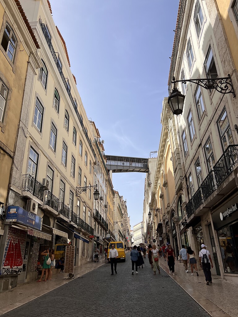 Bustling Lisbon street with historic buildings, pedestrians, and a skybridge connecting two buildings, capturing the vibrant atmosphere of the city.