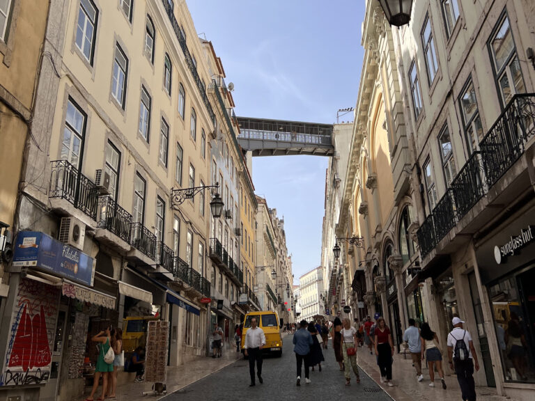 Bustling Lisbon street with historic buildings, pedestrians, and a skybridge connecting two buildings, capturing the vibrant atmosphere of the city.